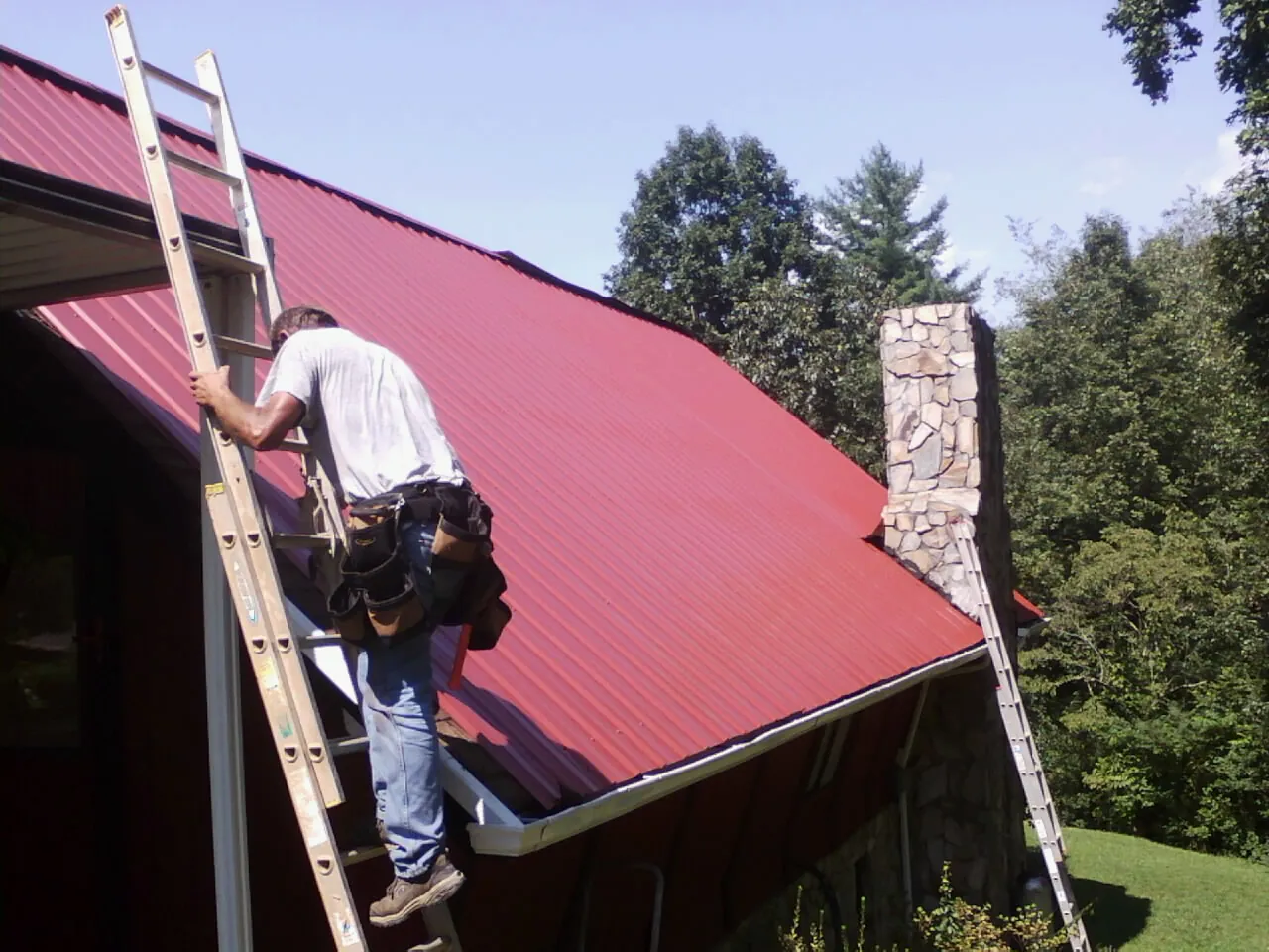 Roofer on a ladder beside a red metal roof and stone chimney — Soffit & Fascia Repair by Roofing Brothers in Mc Lean